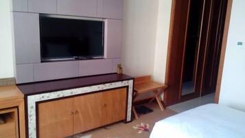 A hotel room with a large flat-screen TV mounted on the wall, a wooden cabinet with drawers, a small wooden chair, and a bed with white bedding.