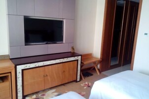 Habitación de hotel con un televisor de pantalla plana grande montado en la pared, un armario de madera con cajones, una pequeña silla de madera y una cama con ropa blanca.