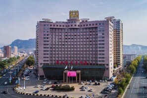A multi-story hotel with a central entrance, surrounded by a parking lot and a busy street. - Junan Gloria Plaza Hotel (Chengde County)
