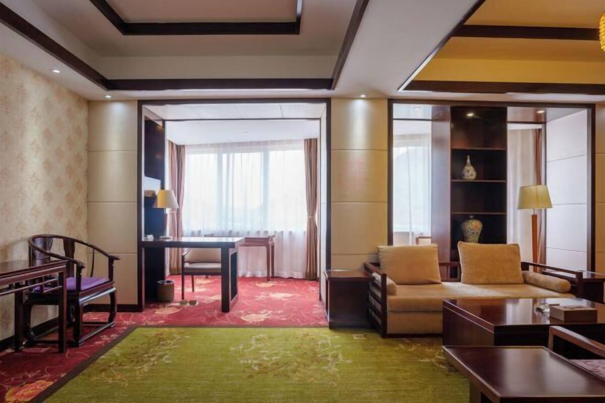 A hotel room with a sofa, a dining table, and a chair. There is a window with curtains and a view of the outside.