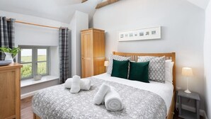 A bedroom with a bed, a window with curtains, a wooden wardrobe, a bedside table with a lamp, and a wall-mounted picture frame. - Cosy Rural Garden Cottage Sea Views Near St Ives (Cornwall)