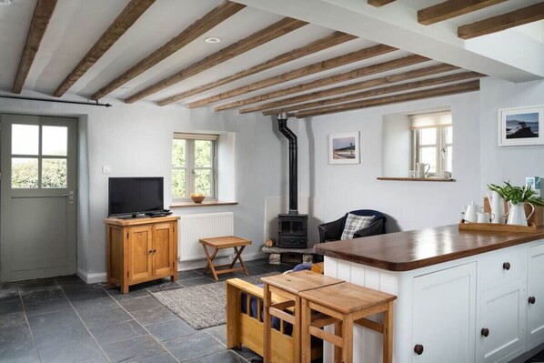 A cozy kitchen with exposed wooden beams, a built-in stove, and a wooden dining table. - Cosy Rural Garden Cottage Sea Views Near St Ives (Cornwall)