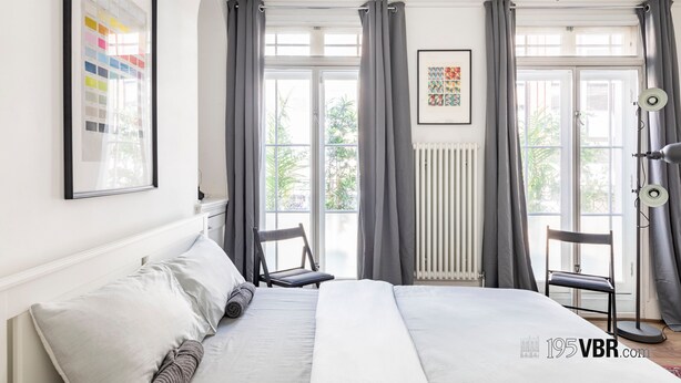 A modern bedroom with a large bed, two chairs, a radiator, and a window with curtains. - The Tachbrook at No. 195: 6-Bed Historic Townhouse | Victoria (London)