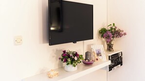 A flat-screen TV mounted on a white wall, a white shelf with a vase of flowers, a candle, and a framed photo. - Apartments Vera (Mljet)