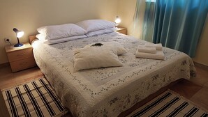 A neatly made bed with a floral-patterned duvet, two bedside lamps, and a striped rug on a tiled floor. - Apartments Vera (Mljet)