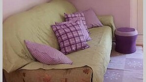 A room with a green sofa, two purple pillows, a purple trash bin, and a window with yellow and purple curtains. - Apartments Vera (Mljet)
