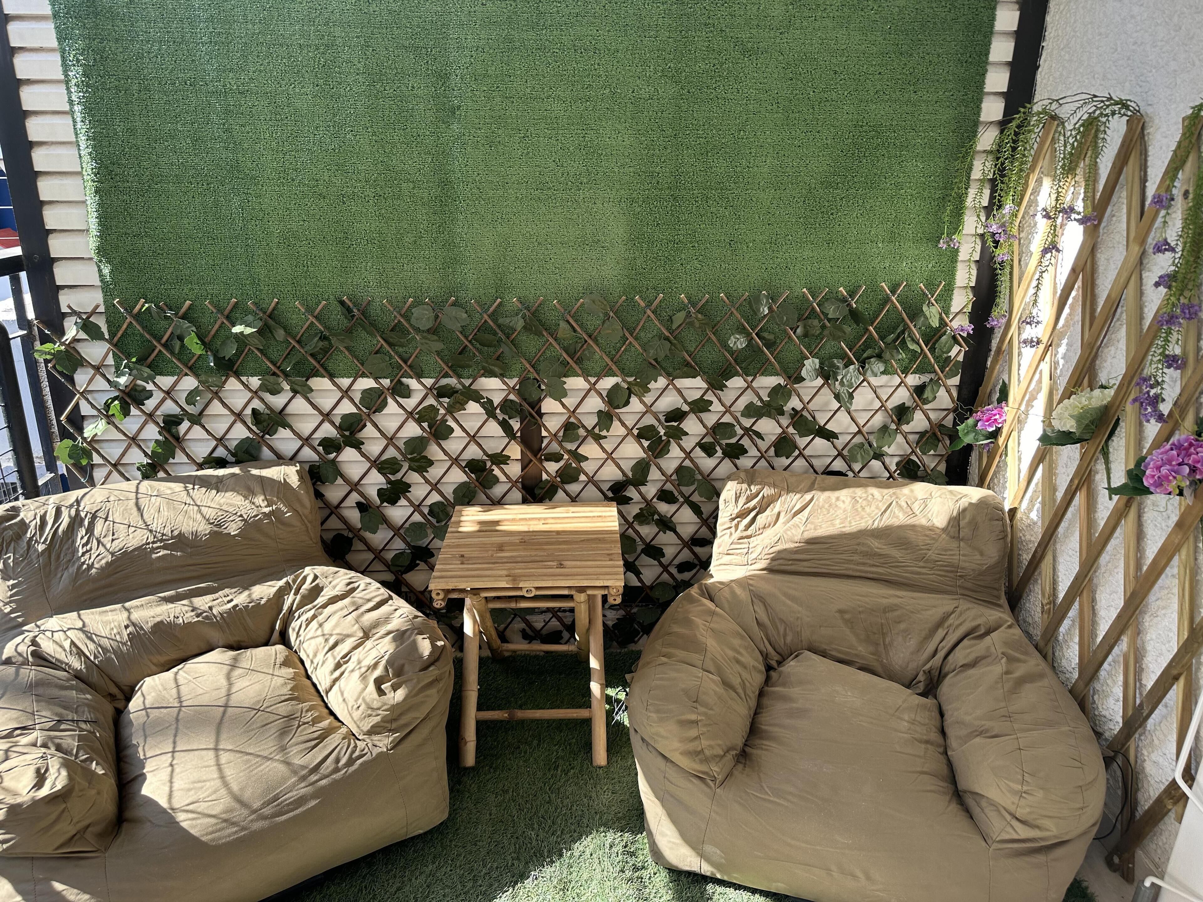 Un espace extérieur accueillant, aménagé avec deux fauteuils beiges confortables et une petite table en bois, le tout entouré d’une clôture en treillis et d’une végétation luxuriante.
