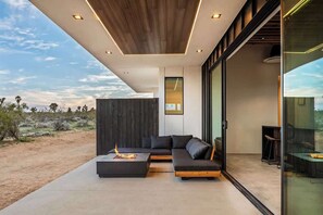 Un salon moderne doté d’un grand canapé d’angle, d’une table basse avec un foyer intégré et d’une porte vitrée donnant sur un espace extérieur.