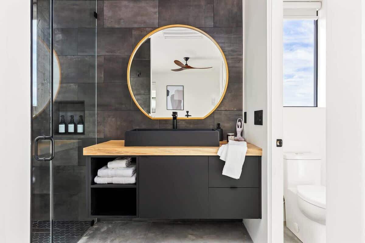 A modern bathroom with a round mirror, dark tiles, a wooden countertop, and a glass shower enclosure.