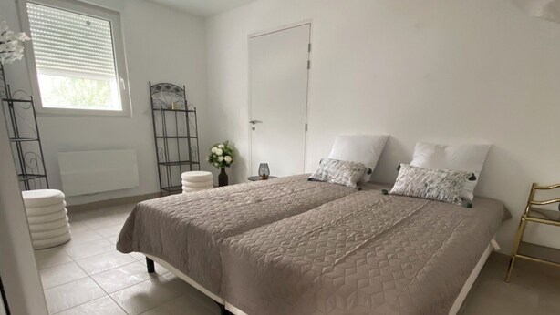 A spacious bedroom with a large bed, a window with blinds, a radiator, and a bedside table with a vase of flowers. - 4 Bedroom Apartment in Camblain-ChÃ¢telain (Camblain-Châtelain)