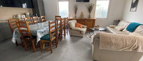 A modern living room with a dining area, a sofa, a TV mounted on the wall, and a kitchen area with cabinets and a countertop.