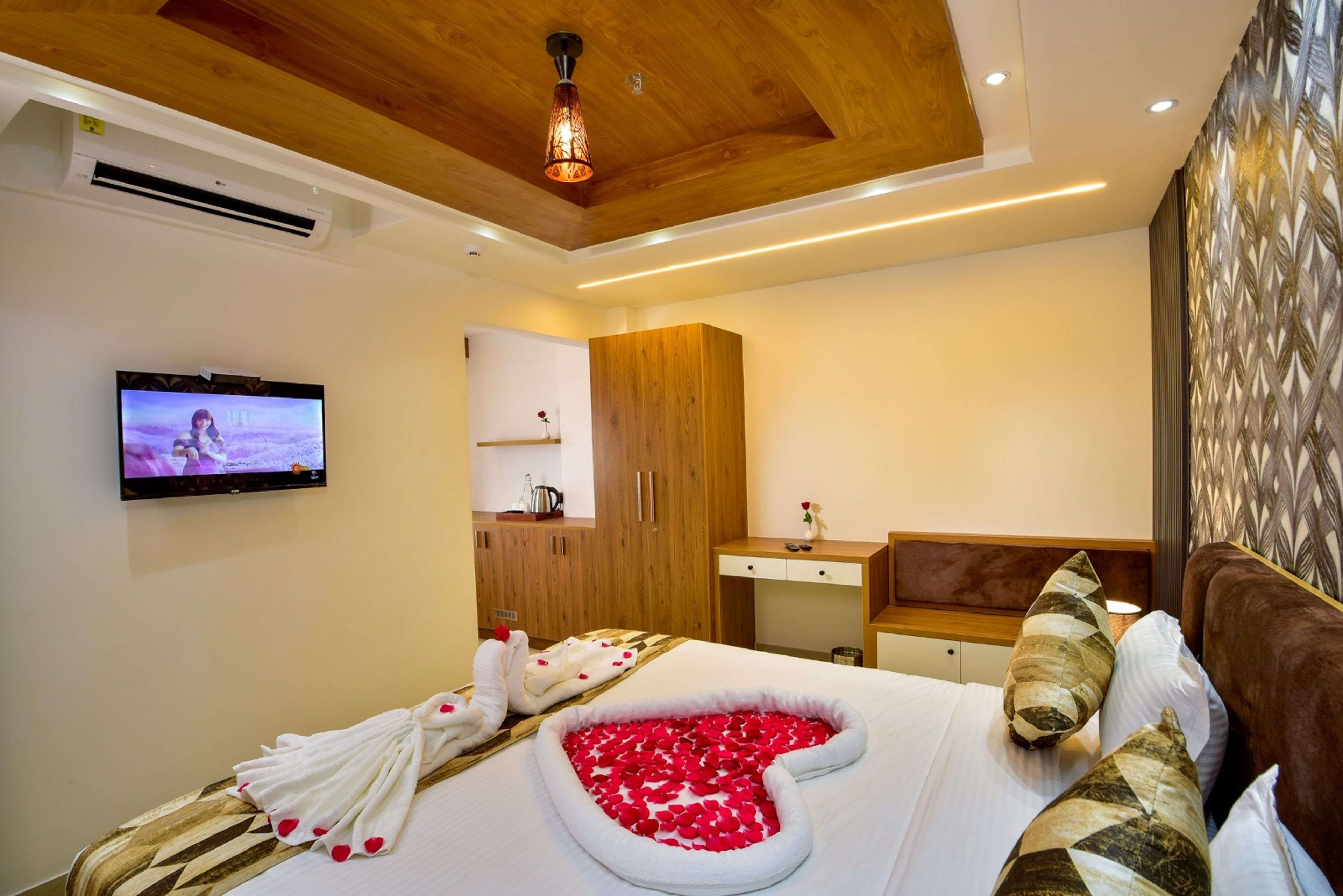 A hotel room with a bed adorned with rose petals, a television, wooden furniture, and a modern ceiling design.