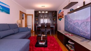 Una sala moderna con un sofá gris, un tapete rojo, un televisor de pantalla plana y una zona de comedor con una mesa y sillas de madera.