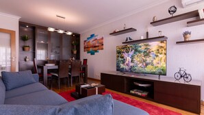 Una sala de estar moderna con un sofá gris, un tapete rojo, un televisor sobre un mueble de madera y un comedor al fondo.