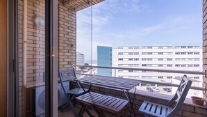 Un balcón con una mesa y sillas, un muro de ladrillo y vista a edificios al fondo.