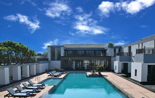A modern hotel with a swimming pool, lounge chairs, and a clear blue sky.