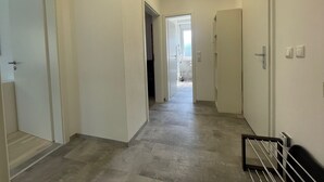 Un couloir avec un plancher de couleur claire, des murs blancs et un luminaire au plafond. Il y a des portes de chaque côté et une main courante en métal sur la droite.