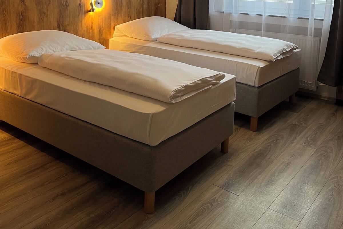 Habitación de hotel con dos camas, suelo de madera, una lámpara fijada a la pared y una ventana con cortinas traslúcidas.