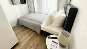 Un dormitorio pequeño y ordenado con una cama, un televisor de pantalla plana montado en la pared, una mesita de noche con un control remoto y una ventana con cortinas traslúcidas.