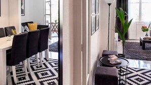 Una sala de estar moderna con zona de comedor, que incluye un tapete de diseño blanco y negro, una mesa de comedor con sillas, una lámpara de pie y una gran planta en maceta.