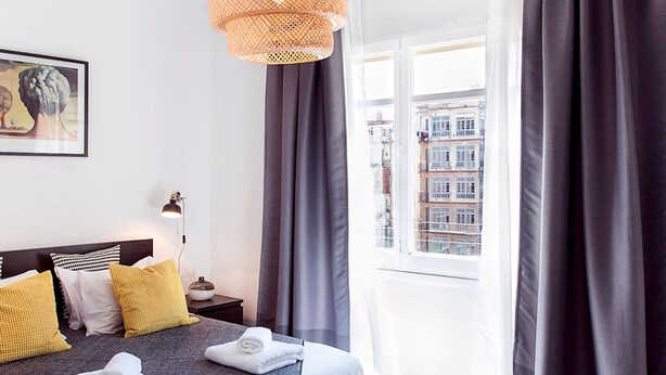 Habitación de hotel moderna con una cama grande, cortinas oscuras, vistas a edificios y una alfombra estampada.