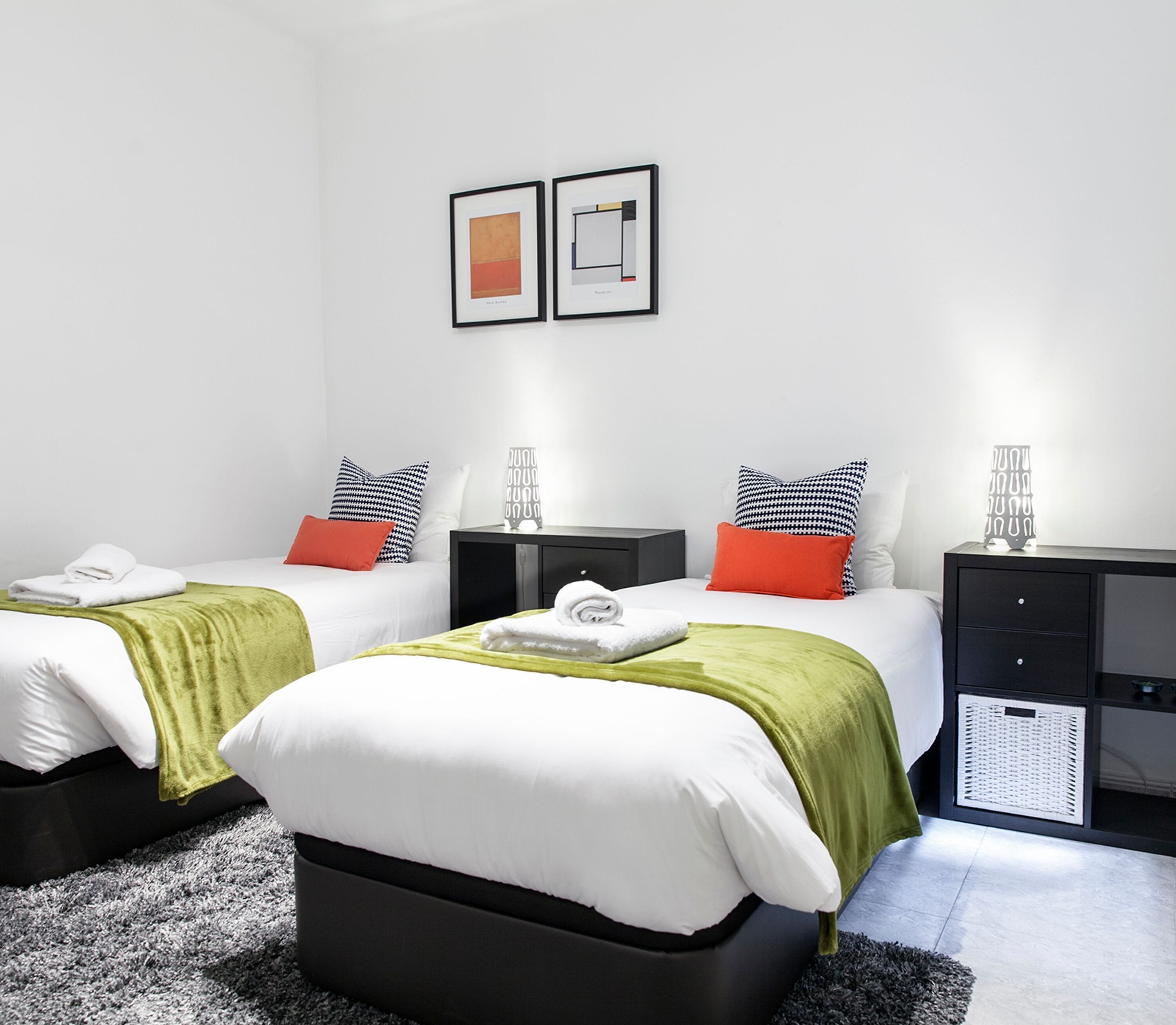 A modern hotel room with two beds, each with a green blanket and white pillows. There are black nightstands with decorative items and a small cabinet with a basket.