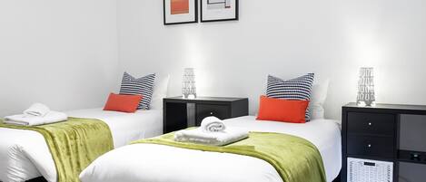 A modern hotel room with two beds, each with a green blanket and white pillows. There are black nightstands with decorative items and a small cabinet with a basket.