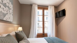 A bedroom with a bed, a TV mounted on the wall, a window with curtains, and a painting on the wall.