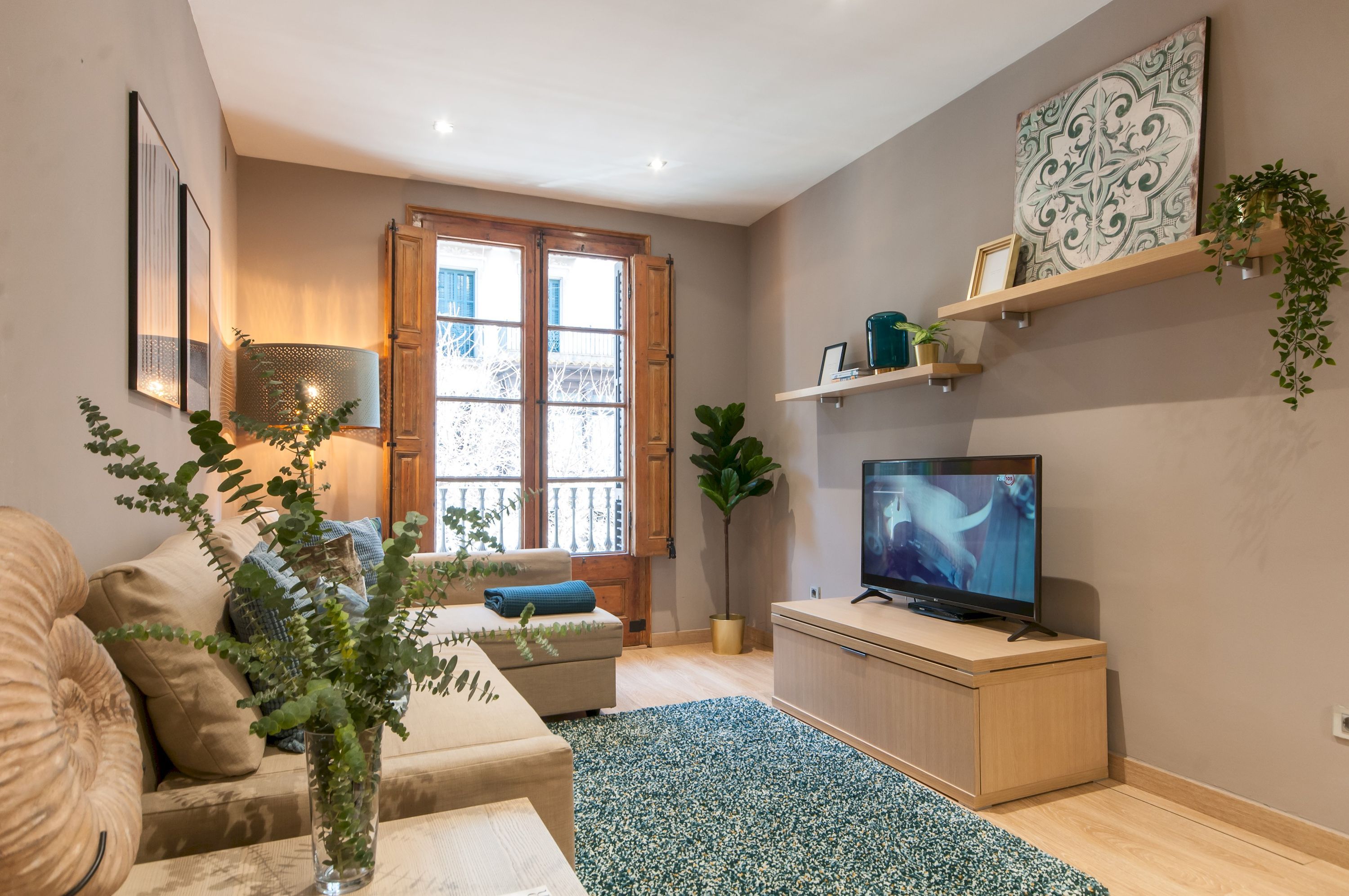 A living room with a sofa, a TV, a coffee table, a potted plant, a window with wooden shutters, and a wall-mounted shelf with decorative items.