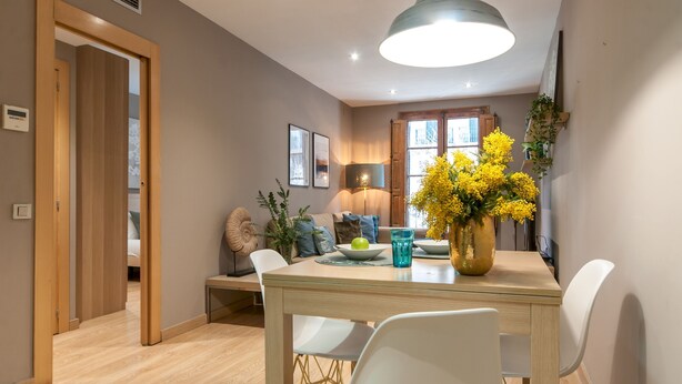 A dining area with a table, chairs, a vase with yellow flowers, a lamp, and framed pictures on the wall.
