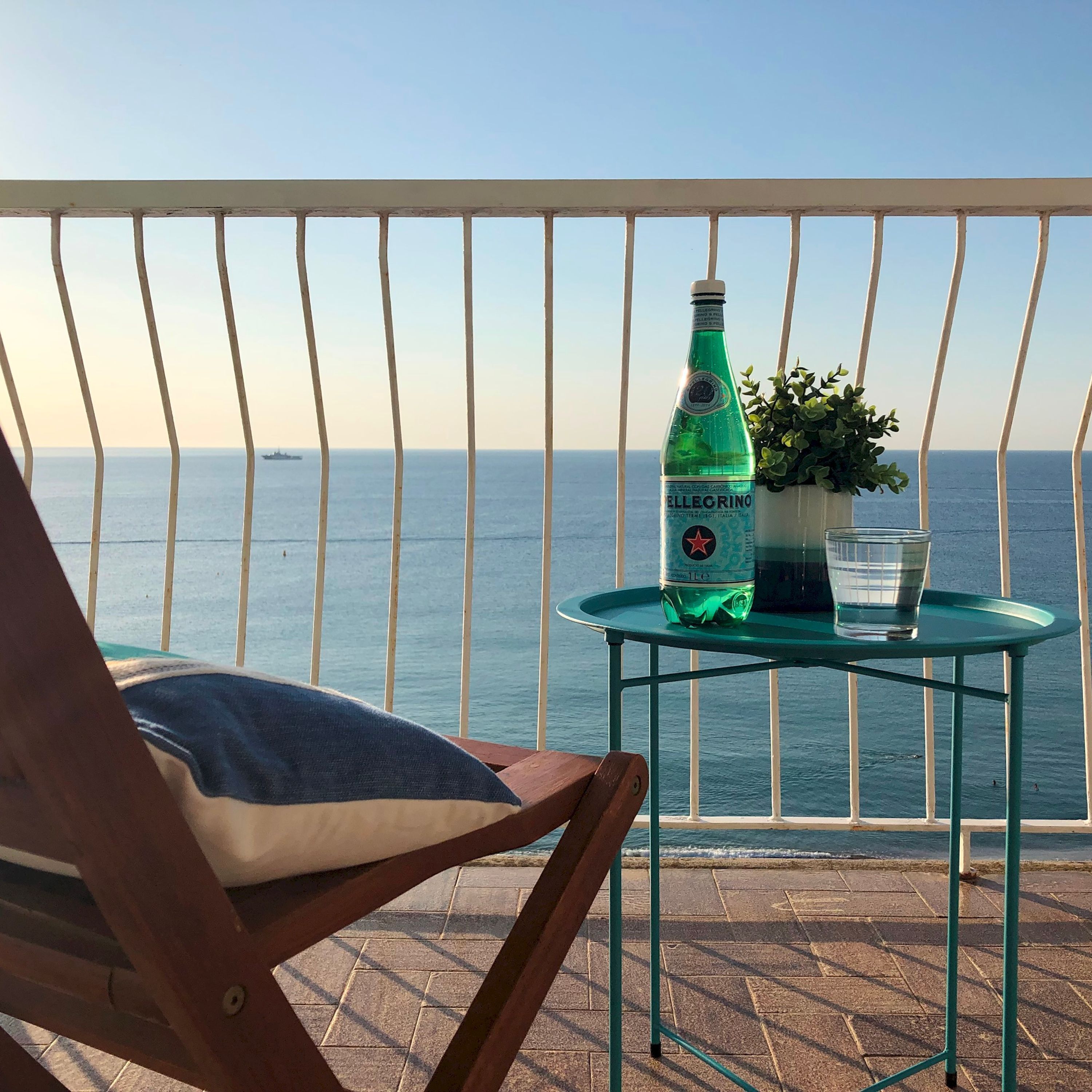 A balcony with a view of the sea, a bottle of El Greino, a glass of water, and a potted plant.