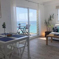 Un salon moderne comprenant un canapé, une table basse et un coin repas avec une table et des chaises. Une grande porte-fenêtre coulissante donne accès à un balcon offrant une vue sur la mer.