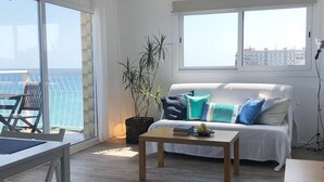A modern living room with a sofa, a coffee table, and a dining area with a table and chairs. There is a large window providing a view of the sea.