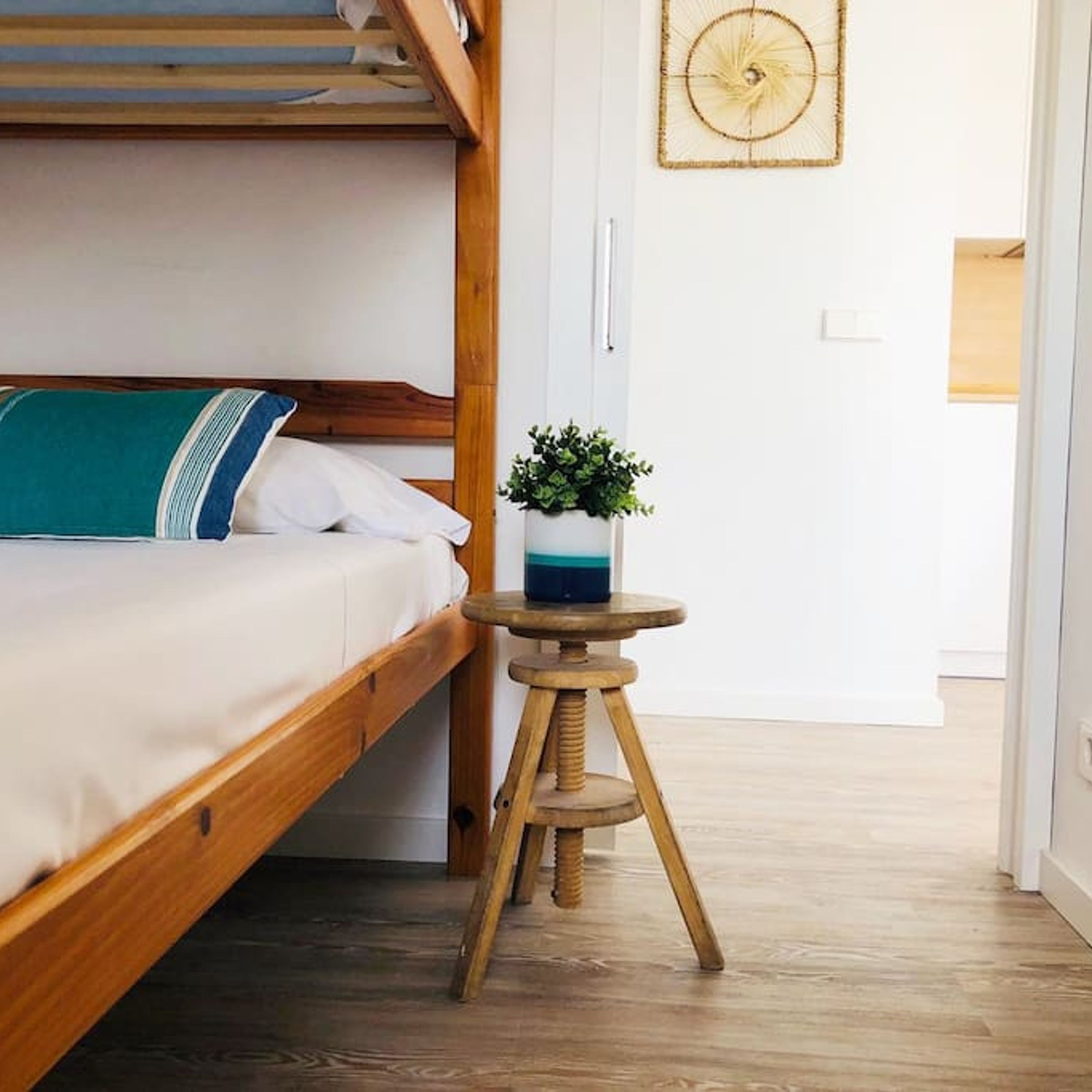 Un lit superposé avec un oreiller turquoise et blanc, un petit tabouret en bois sur lequel est posée une plante, et un mur orné d’un motif circulaire.