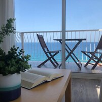 Un balcón con dos sillas de madera mirando al mar, una planta en maceta y un libro abierto sobre una mesa.
