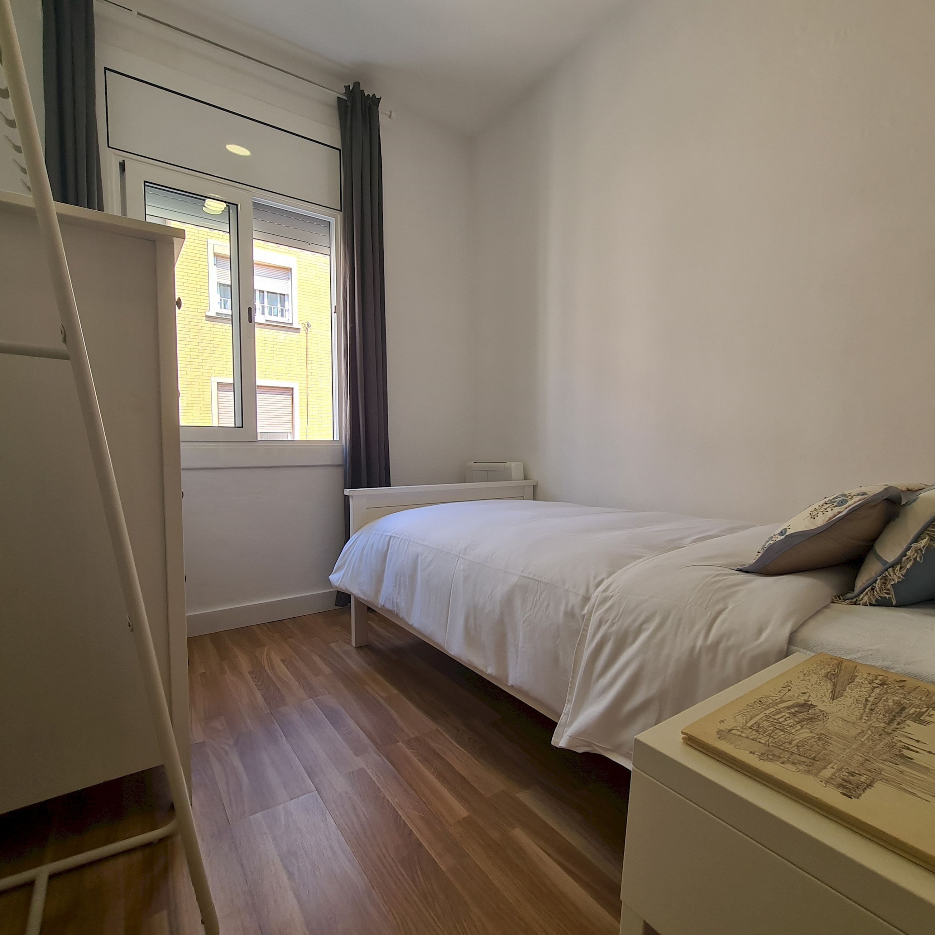 A bedroom with a bed, a bedside table, a window with curtains, and a view of a yellow building.