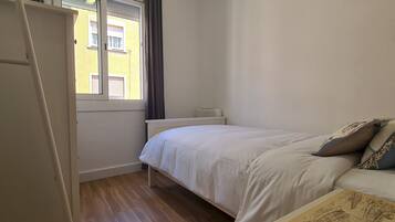 A bedroom with a bed, a bedside table, a window with curtains, and a view of a yellow building.