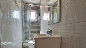 A narrow bathroom with white tiled walls, a glass shower enclosure, a toilet, and a sink with a mirror above it. - Walk to Camp NOU - Wi-fi AC 3 Bed by Myrentalhost (L'Hospitalet de Llobregat)