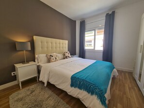 A bedroom with a bed, a nightstand, a lamp, a window with curtains, and a view of the outside.