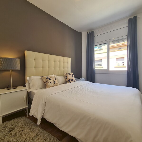 A bedroom with a large bed, a nightstand with a lamp, and a view of buildings through the window.