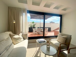 A modern living room with a sofa, armchair, and coffee table. A large sliding glass door leads to an outdoor patio with a table and chairs. - Modern Penthouse With Views Near Paseo De Gracia By Myrentalhost (Barcelona)