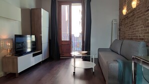 A living room with a TV, sofa, and a small table. There is a balcony door with curtains, a brick wall, and a ceiling with wooden beams. - Catalan Comfort Near Sagrada Familia (Barcelona)