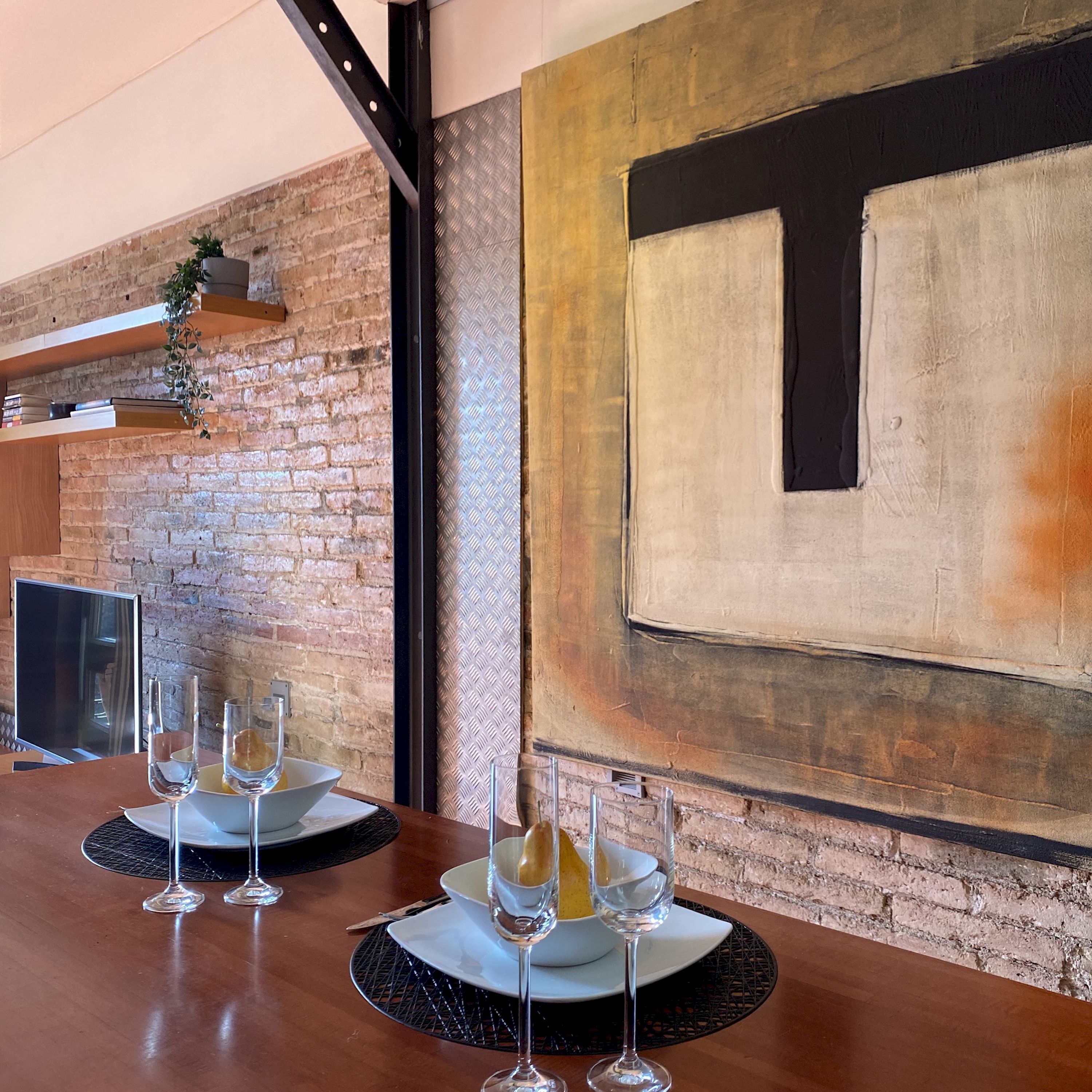 Comedor con pared de ladrillo, un cuadro abstracto grande y una mesa moderna con vasos y platos.