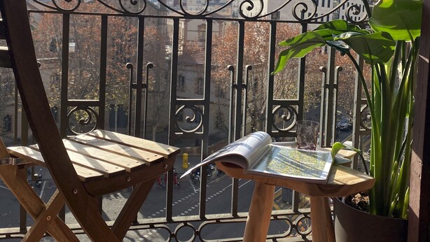 A balcony with a view of a church spire, a potted plant, and a newspaper on a table. - Luminous Designer Loft Style Apt Eixample Views By Myrentalhost (Barcelona)