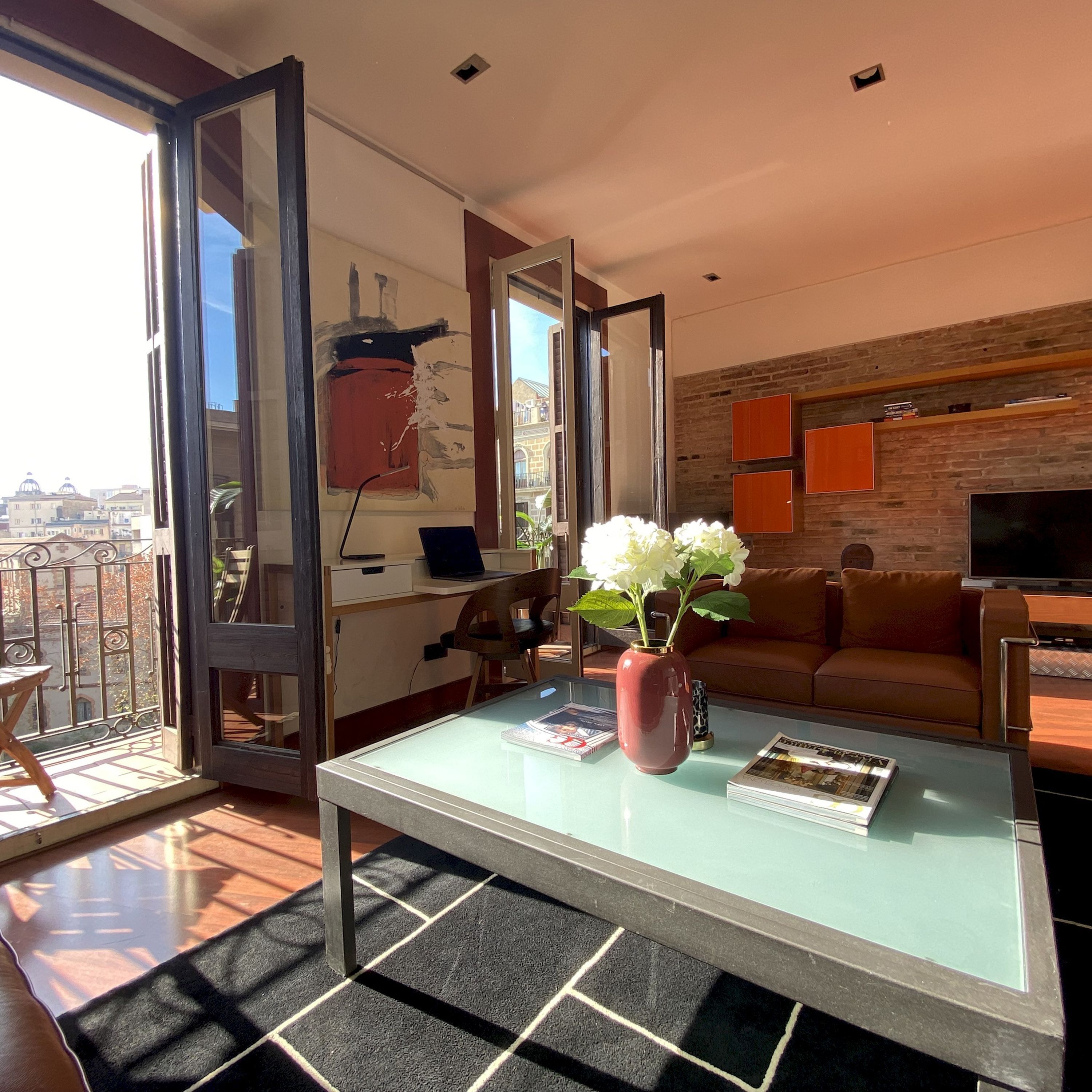 Una sala de estar moderna con una mesa de centro de vidrio, un jarrón rojo con flores blancas, un sofá de cuero y un televisor.