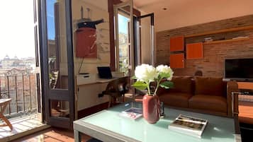 Una sala de estar moderna con una mesa de centro de vidrio, un jarrón rojo con flores blancas, un sofá de cuero y un televisor.