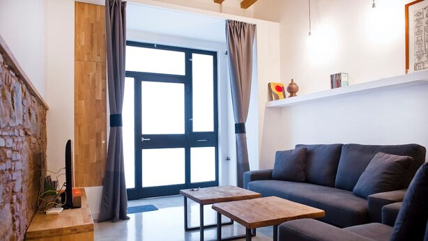 A modern living room with a sofa, two wooden tables, a brick wall, and a large window with curtains. - Unique Architects Apartment IN Trendy Central Poble SEC By Myrentalhost (Barcelona)
