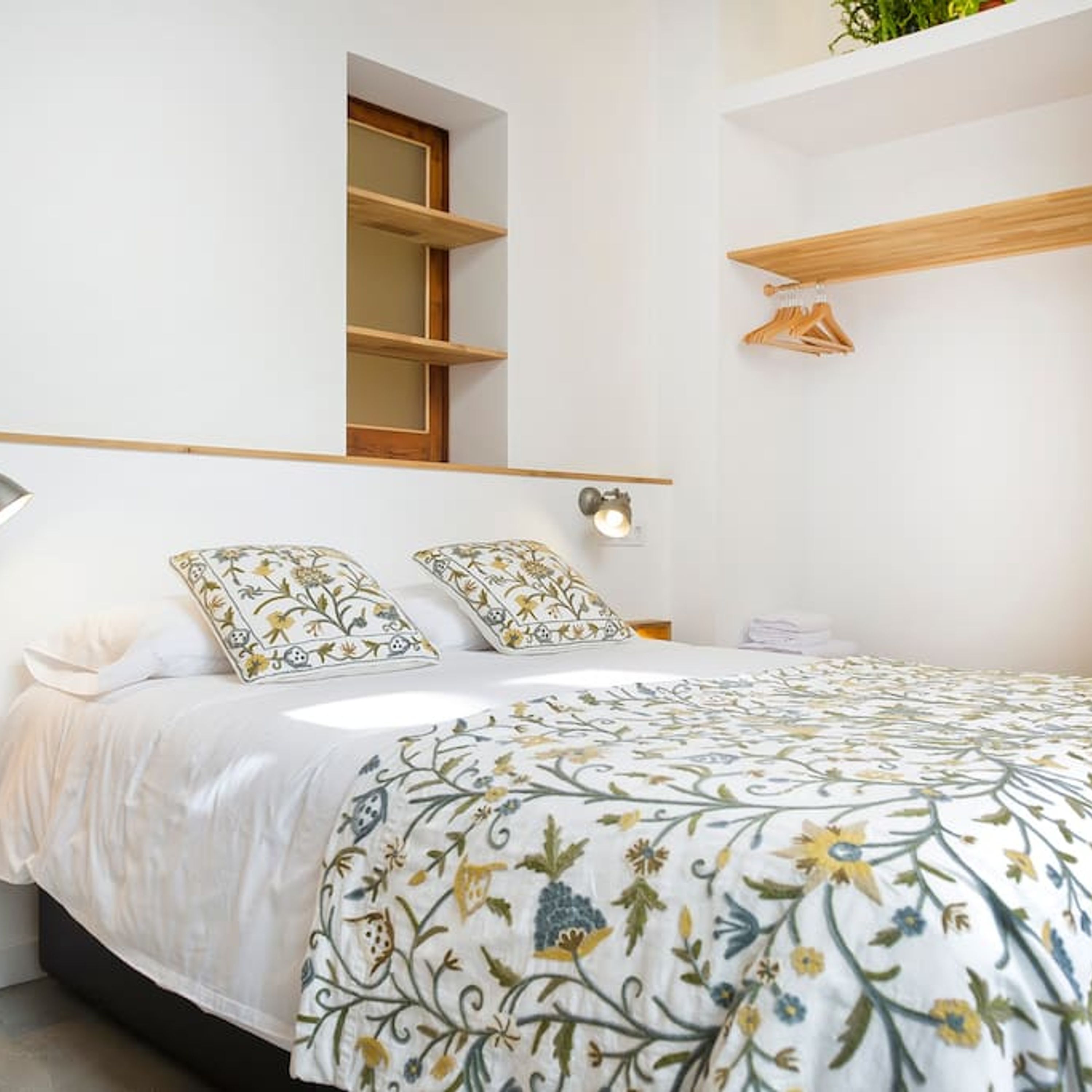 A bedroom with a bed, two pillows, a bedside table, a wall-mounted shelf, and a window.