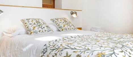 A bedroom with a bed, two pillows, a bedside table, a wall-mounted shelf, and a window.