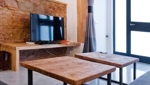 A modern living area with a stone wall, a flat-screen TV, two wooden tables, and a large window with curtains. - Unique Architects Apartment IN Trendy Central Poble SEC By Myrentalhost (Barcelona)